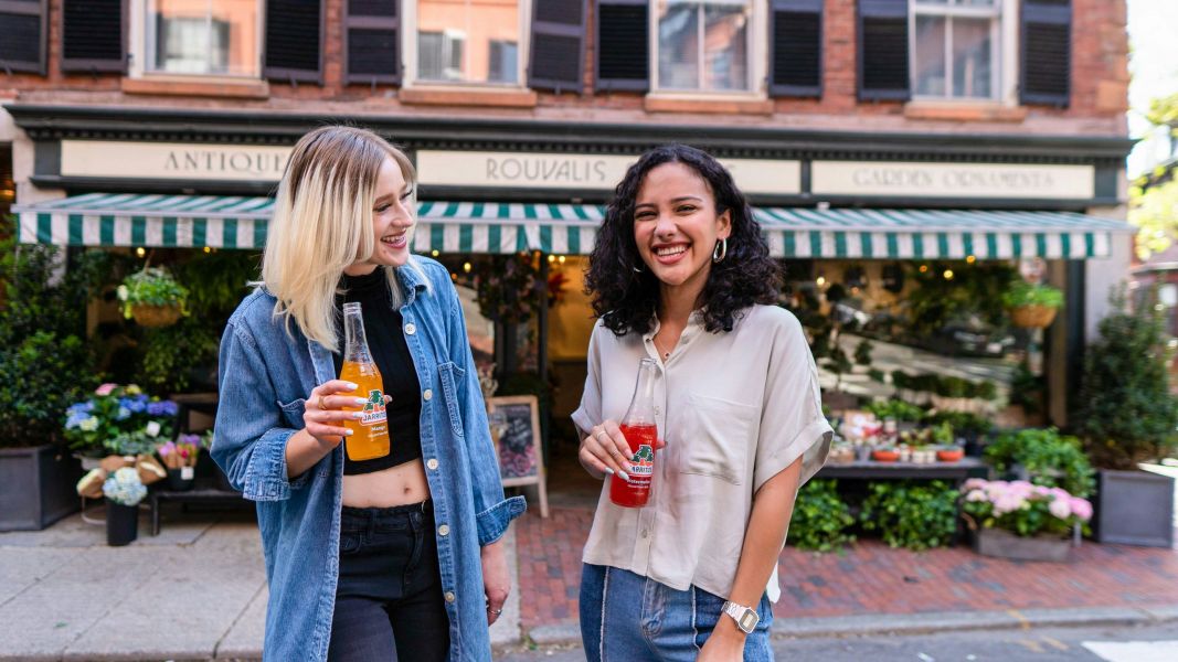 Two people stand outside a shop holding bottled drinks, smiling and conversing with each other on a sunny day.