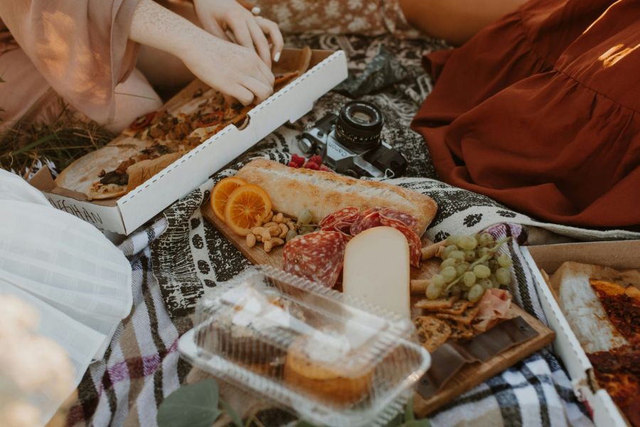 A group of people enjoys a picnic on a blanket with various foods including pizza, orange slices, cheese, nuts, and pastries in a grassy area near The Lodges at Fort Mill.