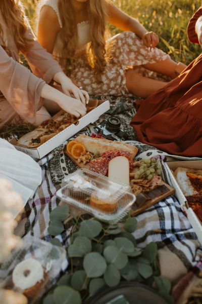 A group of people enjoys a picnic on a blanket with various foods including pizza, orange slices, cheese, nuts, and pastries in a grassy area near The Lodges at Fort Mill.