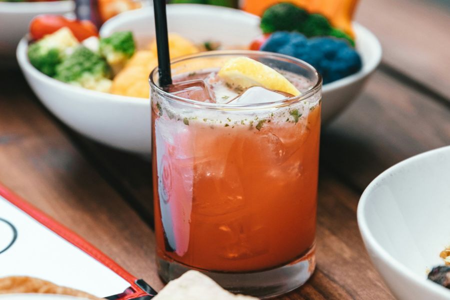 A glass of iced red beverage with a straw and lemon wedge in the foreground, surrounded by bowls of vegetables and dips on a wooden table at The Lodges at Fort Mill.