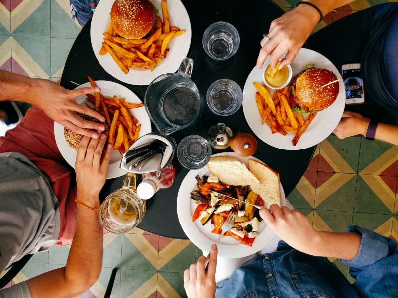 Four people sitting around a table having a meal with burgers, fries, and drinks. One person is also using a smartphone.
