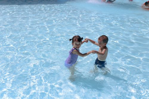 Two children, one girl in a purple swimsuit and one boy in blue shorts, hold hands while playing in a shallow pool at The Lodges at Fort Mill.