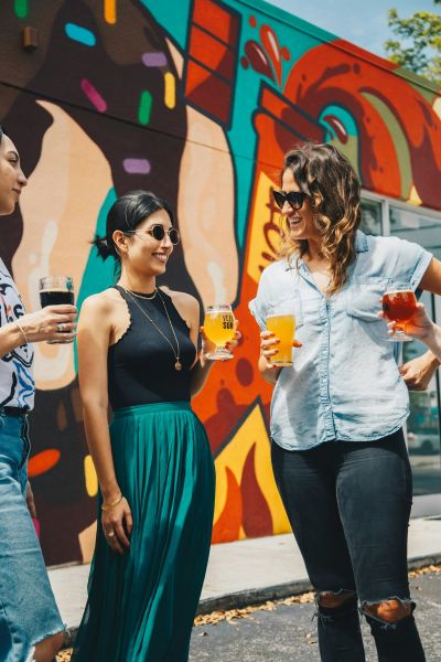 Three people stand outdoors near a colorful mural, smiling and holding drinks in their hands.