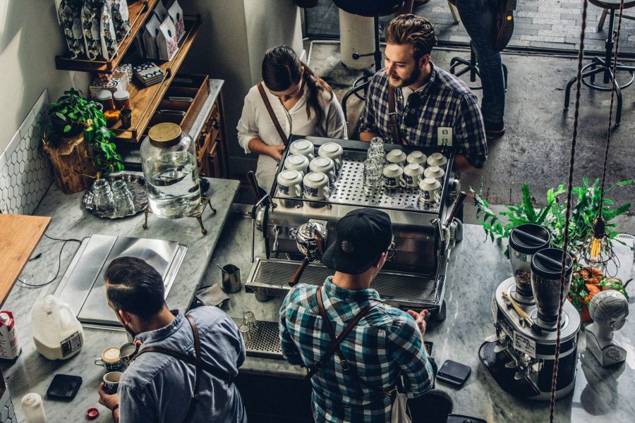 Three people wearing aprons prepare coffee drinks behind a counter in a busy coffee shop while a customer uses her phone nearby.