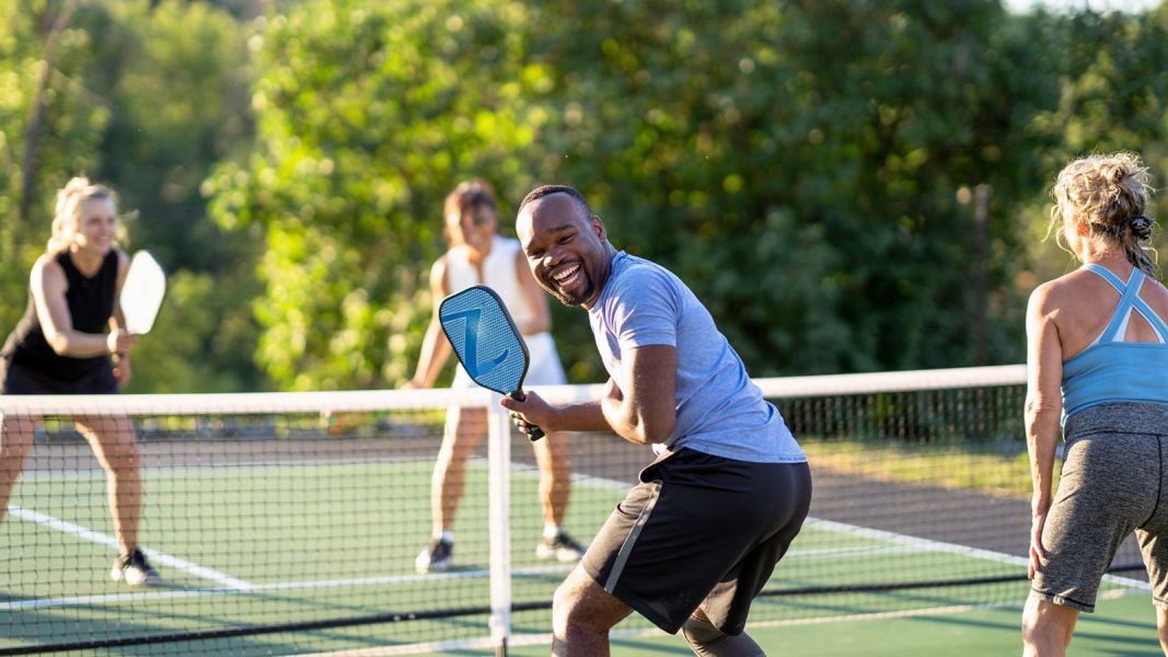 Four people are playing pickleball on an outdoor court at The Lodges at Fort Mill. A man in the foreground is getting ready to hit the ball.
