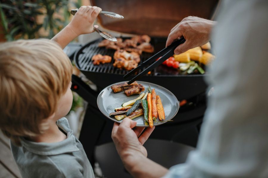 At The Lodges at Fort Mill, an adult and a child are barbecuing vegetables and meat on a grill, with the child holding a plate of grilled vegetables.