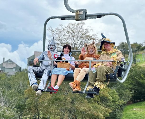 Four people in Wizard of Oz costumes ride a ski lift; one is dressed as the Tin Man, another as Dorothy, the third as the Cowardly Lion, and the fourth as the Scarecrow. It's a whimsical scene you might encounter at Lodges at Fort Mill.