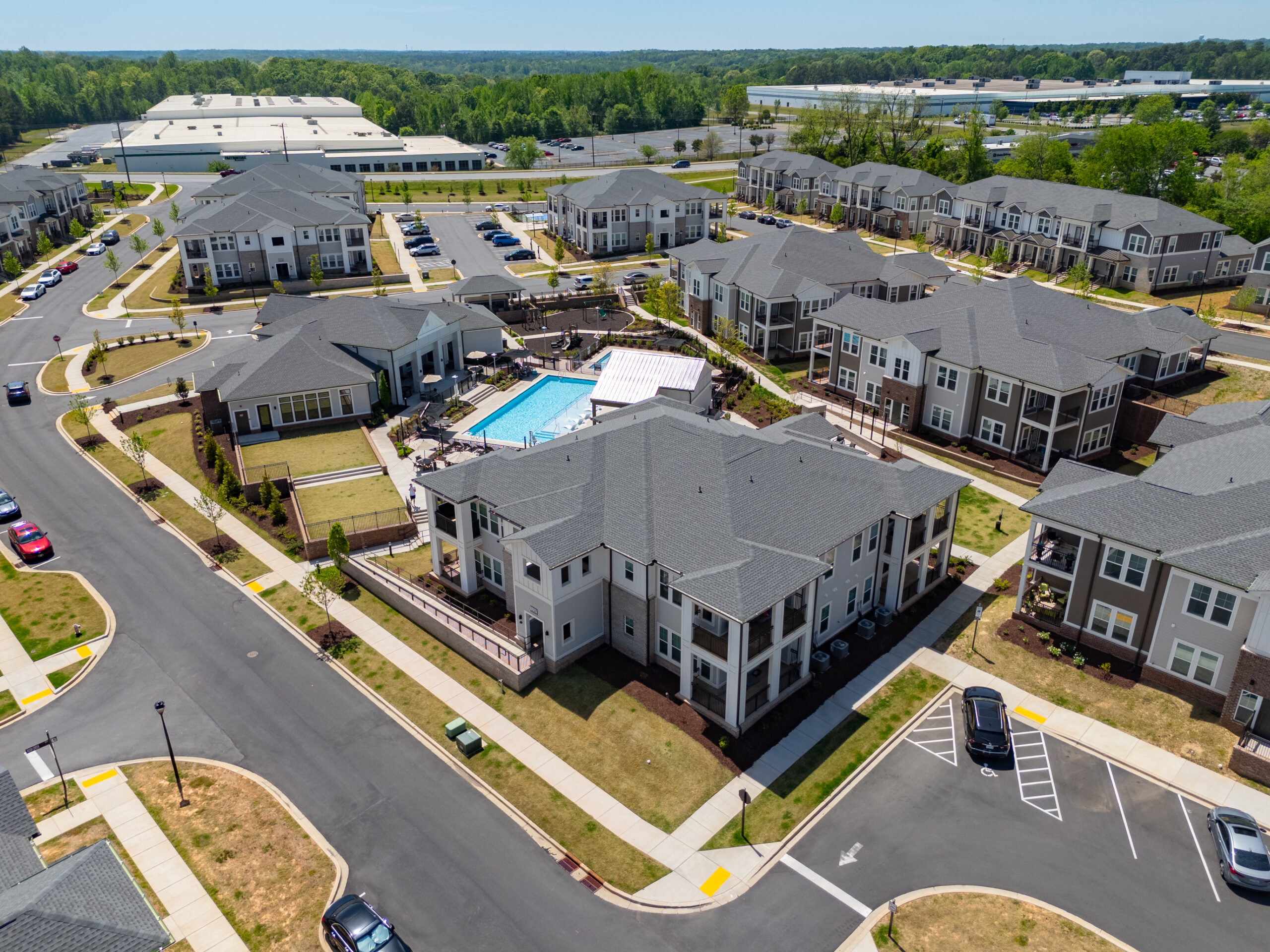 Aerial view of a modern apartment complex with a central swimming pool, parking lots, and landscaped areas on a sunny day.