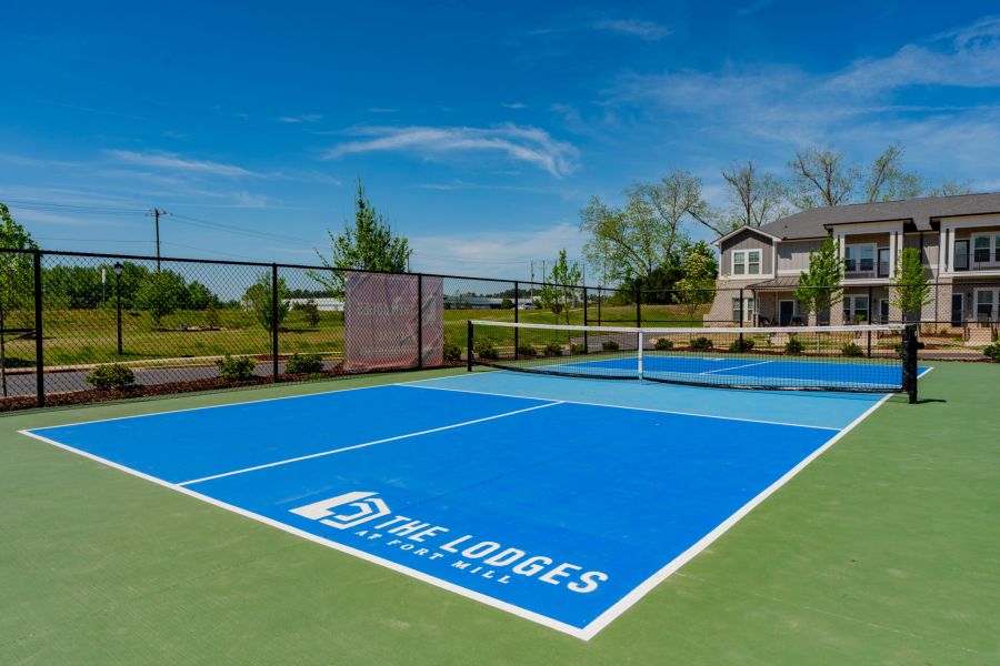 A blue pickleball court with "The Lodges at Fort Mill" logo, surrounded by fencing and greenery, with buildings in the background.
