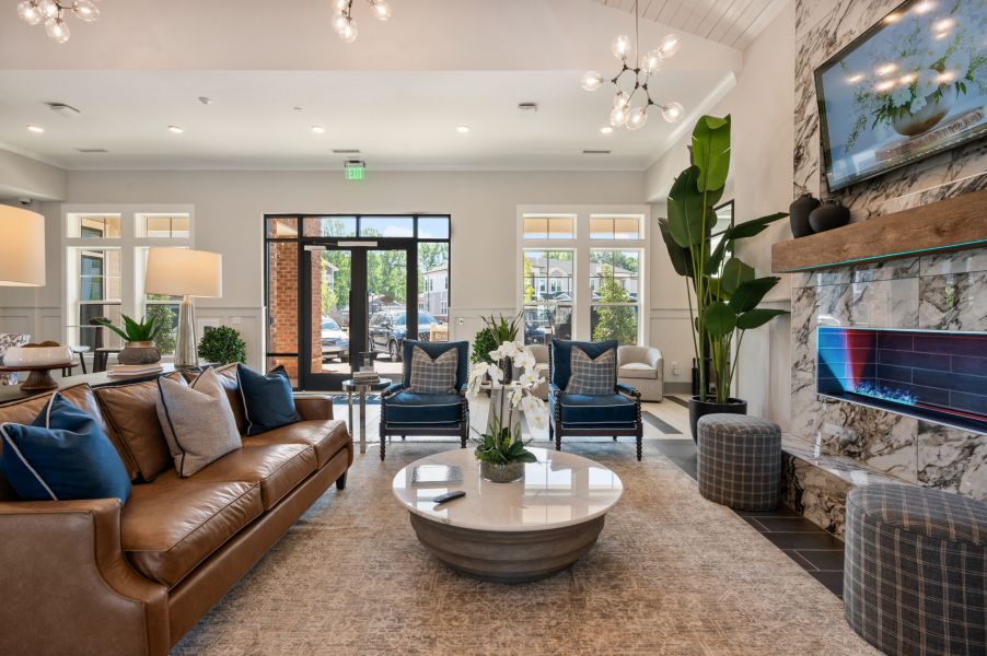 Modern living room with a brown leather sofa, round coffee table, fireplace, TV, and large windows letting in natural light.