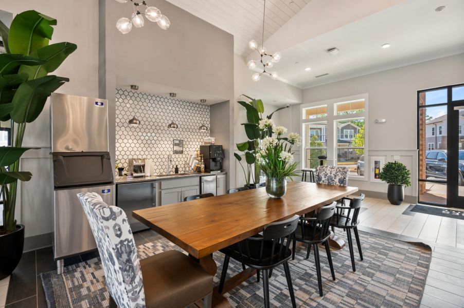 Modern communal kitchen and dining area with a wooden table, black chairs, plants, and natural light from large windows.