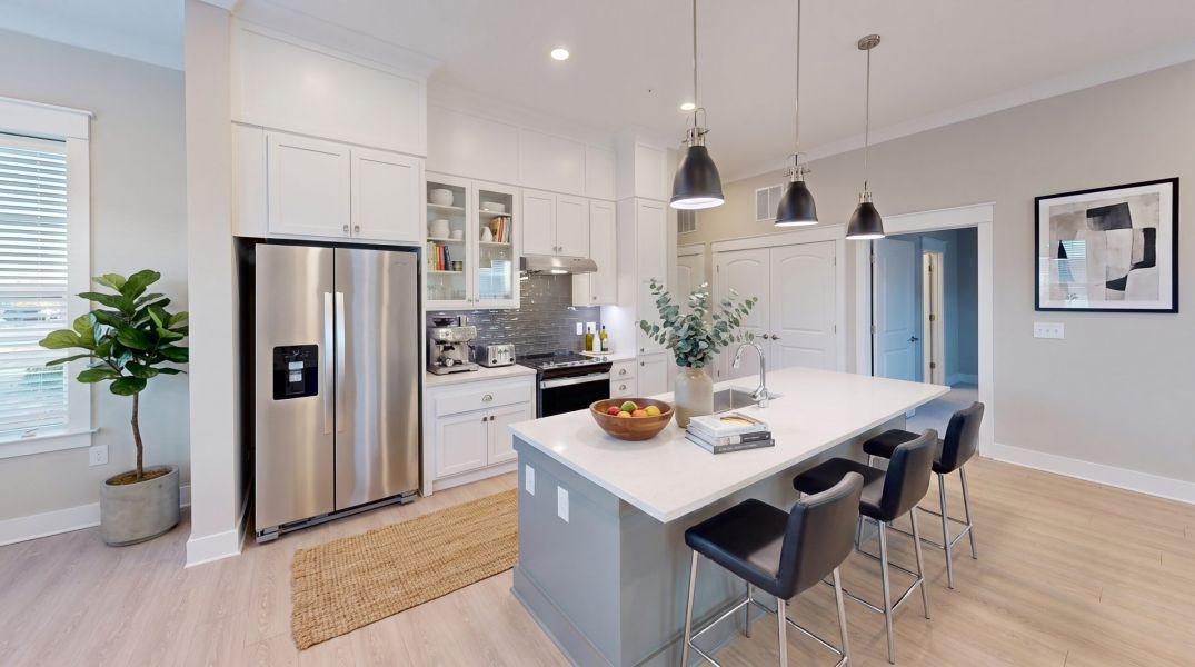 Modern kitchen with white cabinets, stainless steel appliances, island with stools, and hanging lights; plant and framed art on wall.