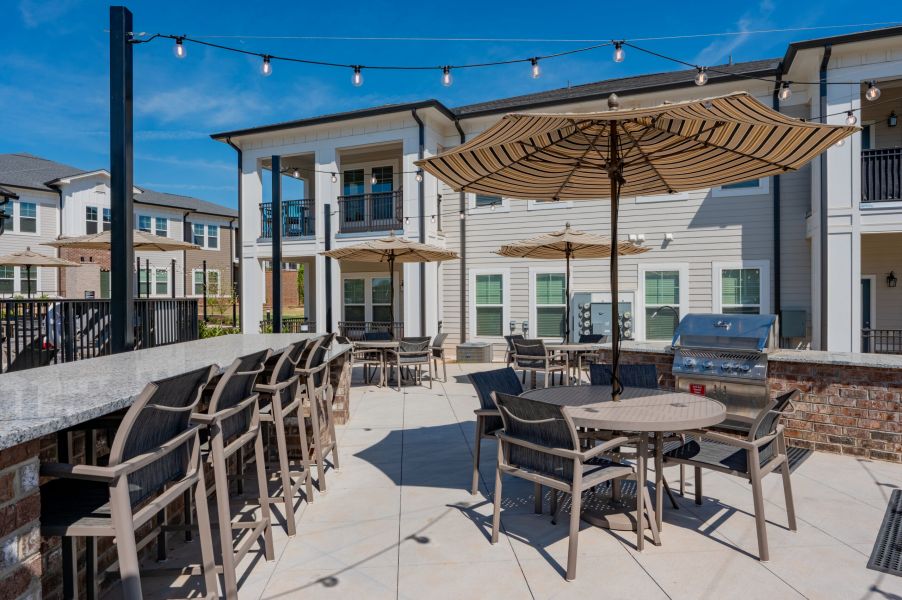 Outdoor patio area with tables, chairs, umbrellas, string lights, and a stainless steel grill in front of apartment buildings.