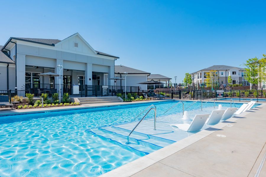 Outdoor swimming pool with in-water lounge chairs, surrounded by modern residential buildings and clear blue sky.