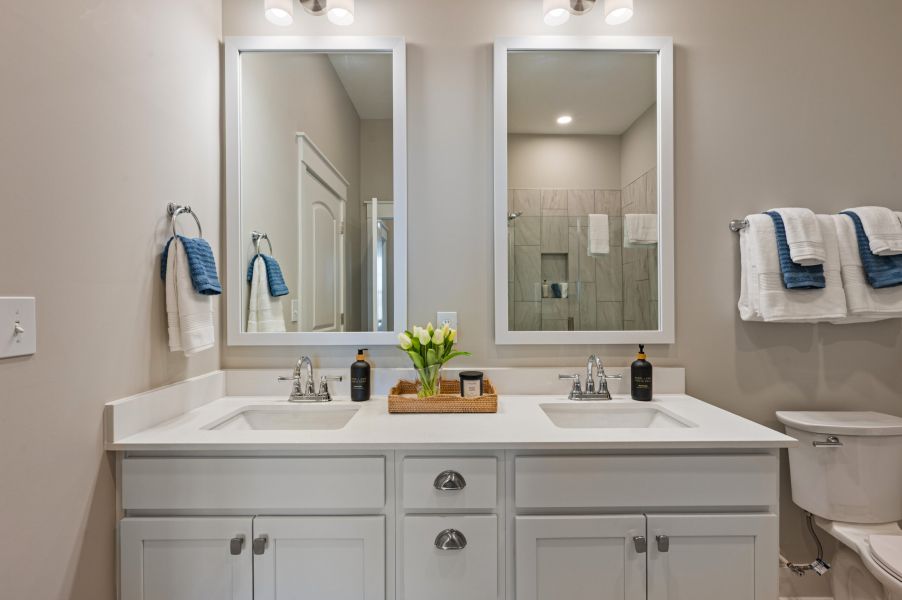 Modern bathroom with double sinks, two mirrors, a tray with flowers and toiletries, blue towels, and a toilet on the right.