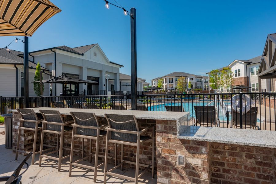 Outdoor seating area with bar stools overlooks a fenced swimming pool and modern apartment buildings under a clear blue sky.