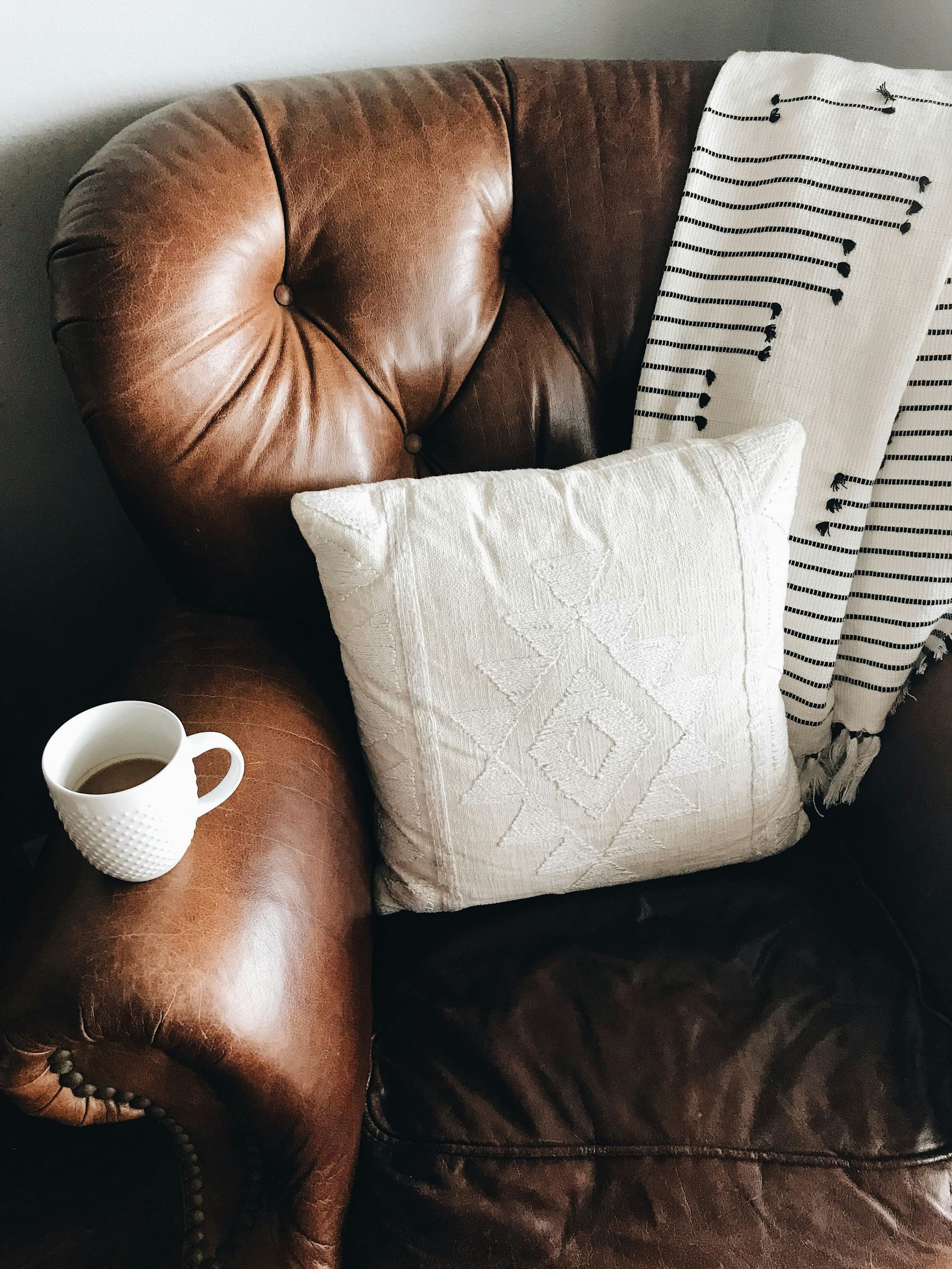 A brown leather armchair with a white throw pillow and a patterned blanket, holding a white mug of coffee on the armrest, perfectly embodies the cozy ambiance of The Lodges at Fort Mill.