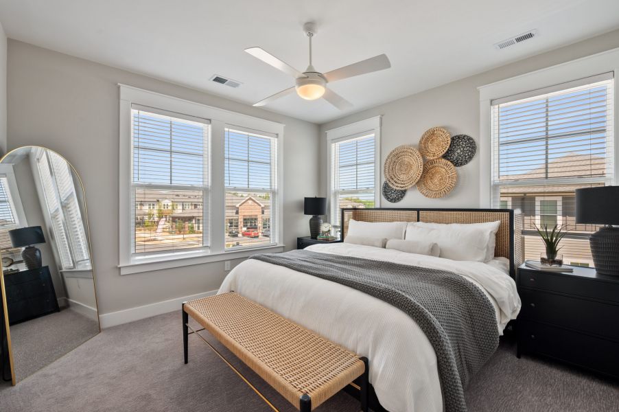 Modern bedroom with a large bed, woven bench, wall decor, two windows, black nightstands, and a floor mirror.