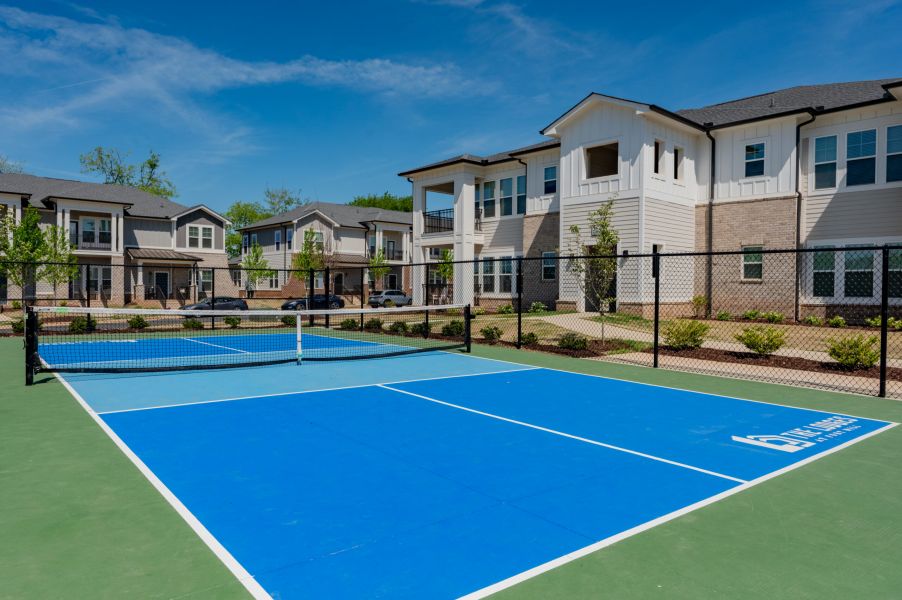 Outdoor blue pickleball court with a net, surrounded by a black fence and modern apartment buildings in the background.