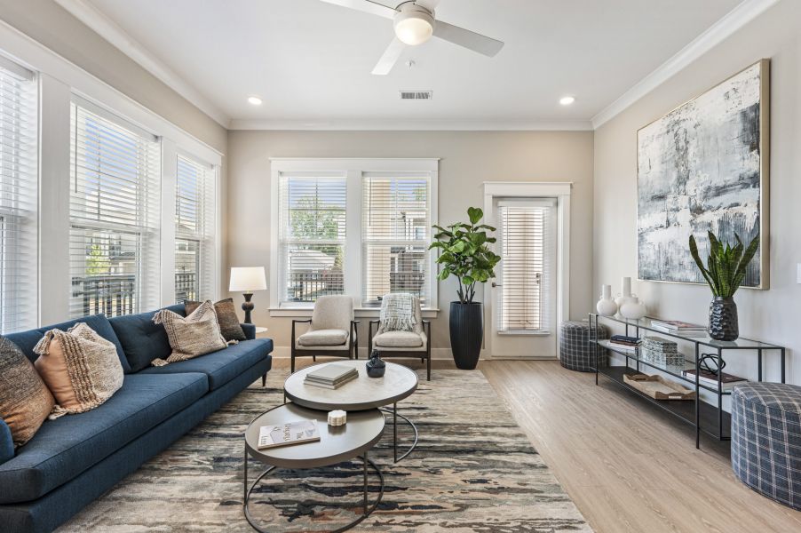 Modern living room with a blue sofa, two chairs, round coffee tables, large artwork, houseplants, and windows letting in natural light.