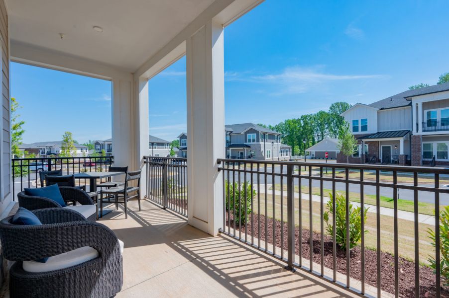 Covered balcony with black railing, outdoor seating, and a view of a suburban street with modern houses on a sunny day.