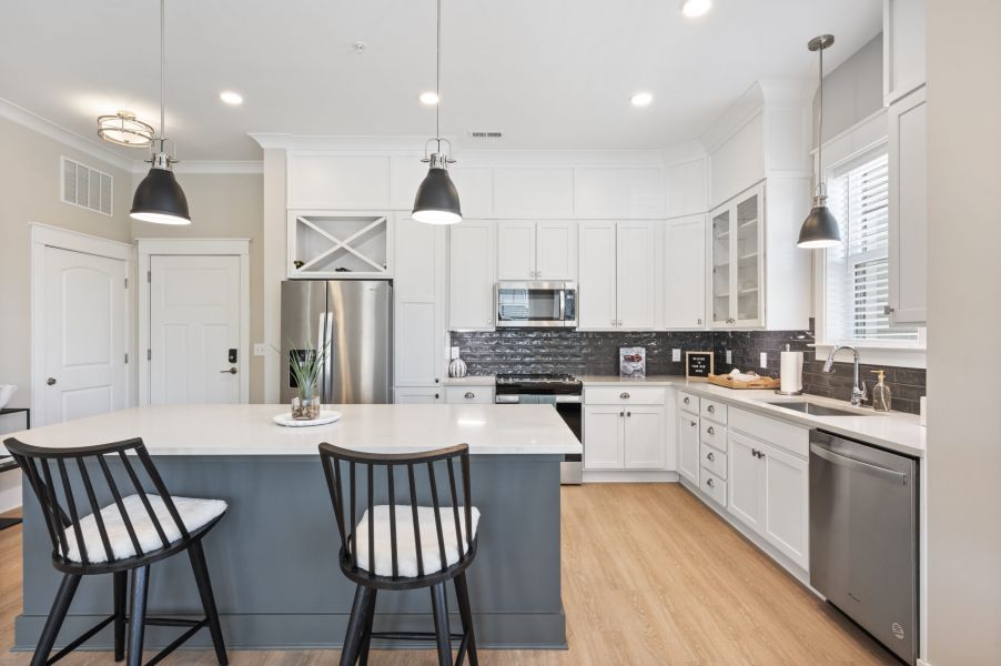 Modern kitchen with white cabinets, stainless steel appliances, a gray island, pendant lights, and two black chairs on wood flooring.