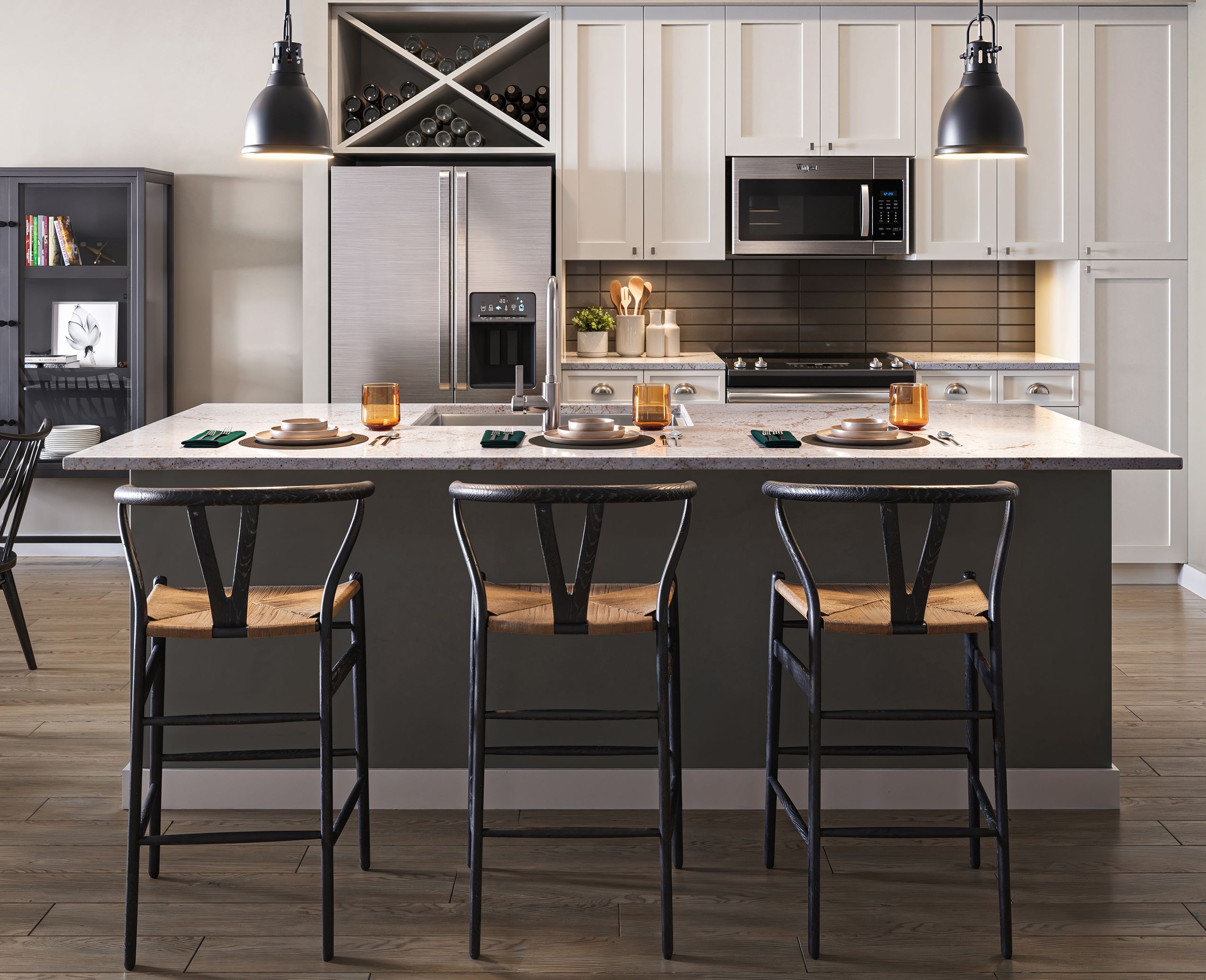 A modern kitchen at The Lodges at Fort Mill features a central island, three bar stools, stainless steel appliances, pendant lights, and a small bookcase with items on display.